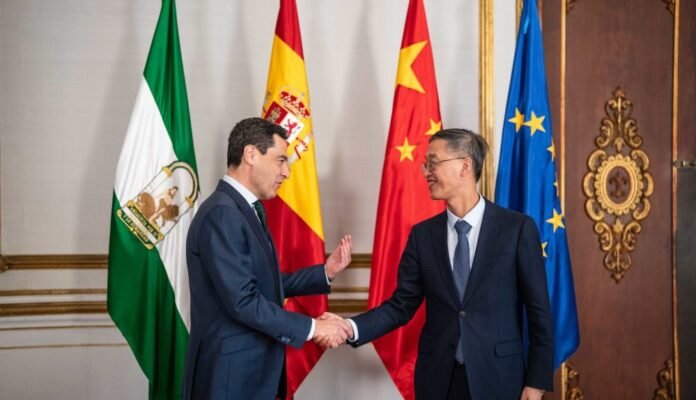 Juanma Moreno, presidente de la Junta de Andalucía, estrecha la mano de un representante chino ante banderas de Andalucía, España, China y la Unión Europea.