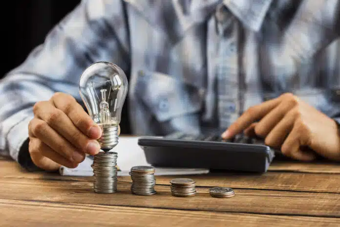 Manos sosteniendo bombilla LED junto a pilas de monedas sobre mesa de madera, simbolizando ahorro energético y financiero