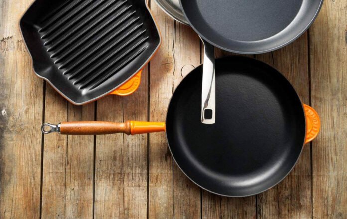 Variedad de sartenes y utensilios de cocina sobre superficie de madera, incluyendo sartén antiadherente, plancha grill y cacerola.