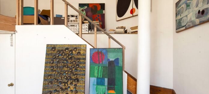 Loft de artista con múltiples pinturas abstractas, escalera de madera, y estantería llena de libros y equipo de sonido, mostrando un espacio creativo ecléctico y lleno de color.