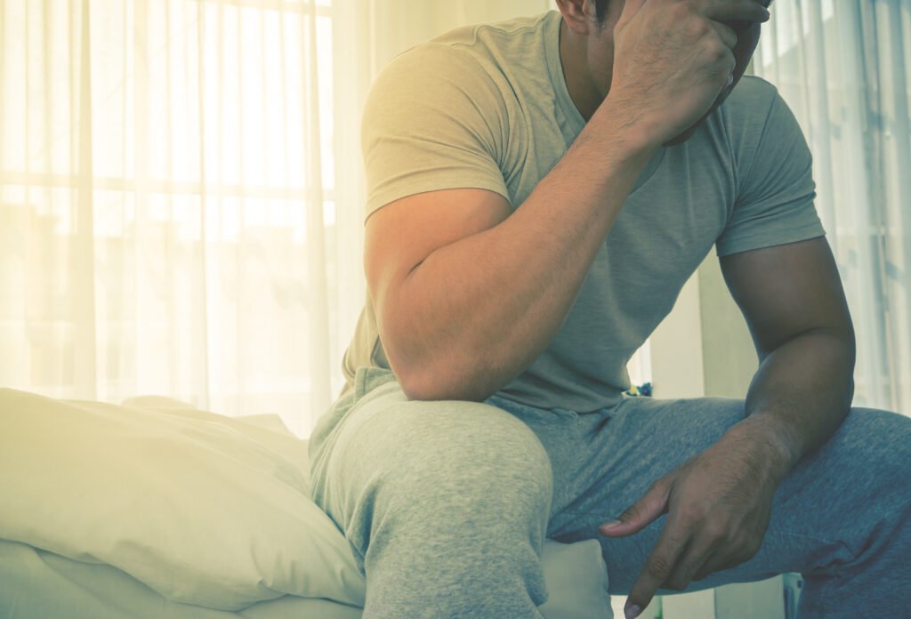 Hombre sentado en el borde de una cama, sosteniendo su cabeza y mostrando signos de incomodidad en la zona pélvica